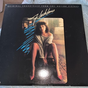 May include: A vintage vinyl record album cover for the movie "Flashdance." The cover is black with the movie title in a stylized font and a woman in a gray sweater and red heels. Blue diagonal lines accent the design.