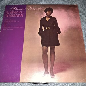 Puede incluir: Portada del álbum "I'll Never Fall in Love Again" de Dionne Warwick. El álbum presenta una foto de Warwick con un vestido negro y una hebilla dorada. La portada del álbum tiene un esquema de color púrpura y melocotón con el título del álbum y los títulos de las canciones.