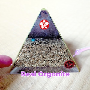May include: A pyramid-shaped orgonite with a clear resin exterior. The interior is filled with small, brown, metallic pieces. The top has a red floral symbol and the text "Real Orgonite" in pink.