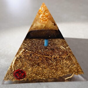 May include: A clear resin pyramid with gold leaf and a blue rod. The pyramid has a layer of dark brown material and a red seal at the base. The pyramid is on a gray surface.