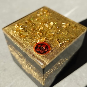 May include: A square, decorative resin box with gold leaf inclusions. The top surface is covered in gold flakes and a red and black emblem. The sides are dark with gold accents, creating a luxurious, eye-catching design.