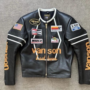 May include: Black leather motorcycle jacket with multiple patches, including the American flag, "Vanson Leathers," and "Champion." White stripes accent the sleeves. The jacket features the text "Vanson" in orange on the sleeves and chest.