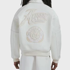 Könnte beinhalten: Cremefarbene Bomberjacke mit dem gestickten Logo "Avirex New York" auf dem Rücken. Die Jacke hat einen gerippten Saum und Bündchen sowie Mesh-Details an den Ärmeln. Auf dem Rücken ist auch ein kreisförmiges Emblem gestickt.