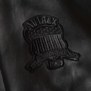 Puede incluir: Primer plano de una chaqueta de cuero negra con un emblema cosido. El emblema presenta la palabra "AVIREX" sobre un dise&ntilde;o de escudo, con "ESTABLISHED NYC 1975" debajo. La chaqueta tiene una superficie texturizada.