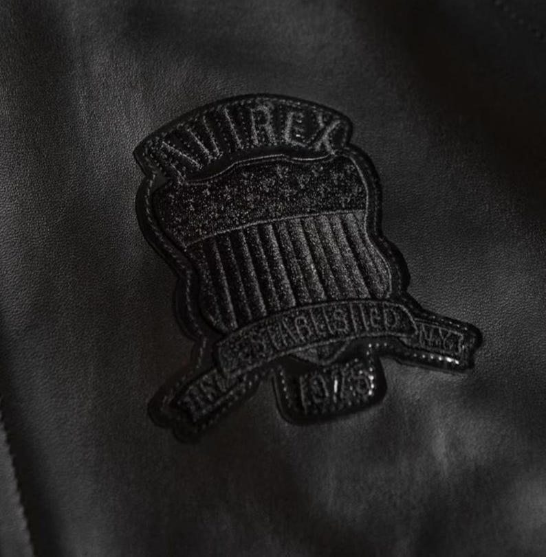 Puede incluir: Primer plano de una chaqueta de cuero negra con un emblema cosido. El emblema presenta la palabra "AVIREX" sobre un dise&ntilde;o de escudo, con "ESTABLISHED NYC 1975" debajo. La chaqueta tiene una superficie texturizada.