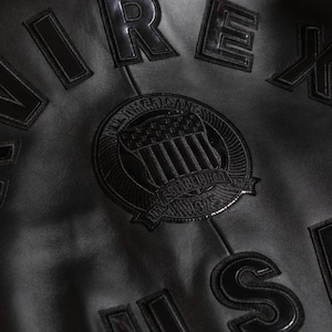 Puede incluir: Primer plano de una chaqueta de cuero negra con letras en relieve y un emblema circular. El emblema presenta un dise&ntilde;o de escudo con las palabras "American" y "Established 1975". La chaqueta tiene una superficie texturizada.