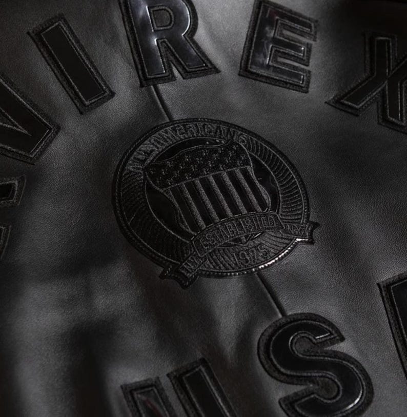 Puede incluir: Primer plano de una chaqueta de cuero negra con letras en relieve y un emblema circular. El emblema presenta un dise&ntilde;o de escudo con las palabras "American" y "Established 1975". La chaqueta tiene una superficie texturizada.