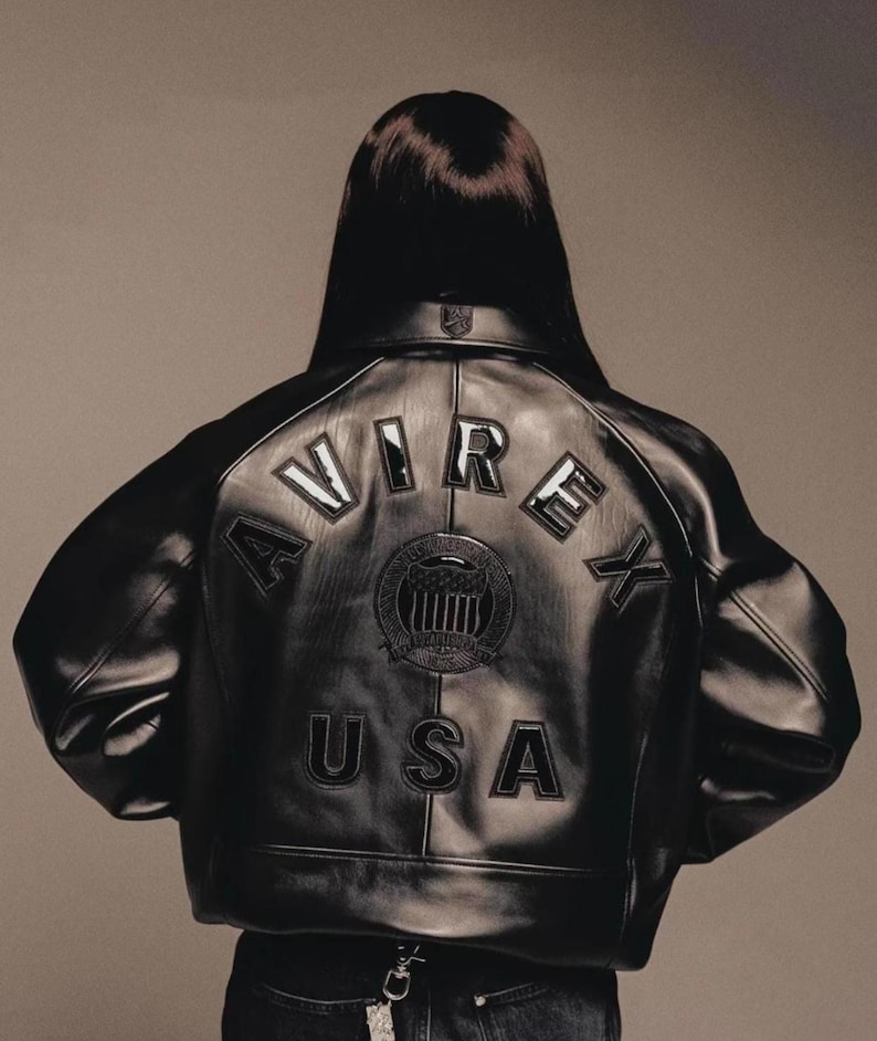 Puede incluir: Chaqueta de cuero negra con la palabra "AVIREX" arqueada sobre los hombros y "USA" debajo de un emblema circular. La chaqueta tiene un estilo bomber cl&aacute;sico con cuello y pu&ntilde;os acanalados. La chaqueta la lleva una persona con pelo largo y oscuro.