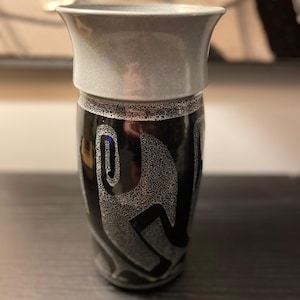 May include: A decorative vase with a flared top and a black body. The vase features a silver-colored rim and a repeating white abstract design. The vase is made of ceramic or glass and is suitable for displaying flowers or as a decorative accent.