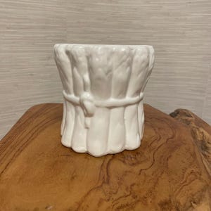 Może przedstawiać: Biała ceramiczna donica w kształcie wiązki pszenicy związanej sznurem. Donica ma teksturowaną powierzchnię i szeroki otwór. Projekt jest szczegółowy, a kolor to jasna biel. Donica stoi na drewnianej powierzchni.