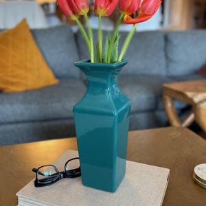 Puede incluir: Un jarrón de cerámica turquesa con tulipanes rojos con tallos verdes. El jarrón está sobre un libro con gafas de montura negra. La escena está ambientada en una mesa de centro de madera, con un sofá gris y un cojín amarillo al fondo.