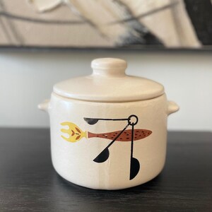 May include: Cream-colored ceramic jar with lid. The jar features a whimsical design of a fork and measuring spoons in yellow, brown, and black. The jar has two small handles and is sitting on a dark surface.