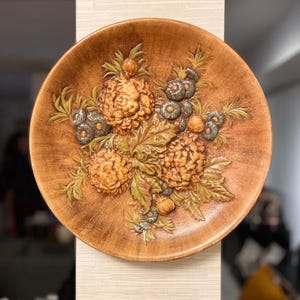 Puede incluir: Un plato decorativo redondo y marrón con un diseño floral en relieve. Las flores son en tonos naranja y amarillo, con hojas verdes y acentos azul-verdes. El plato está montado en una pared de color neutro.