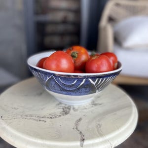 Könnte beinhalten: Eine Keramikschale mit weißem Innenraum und dunkelblauer Außenseite, die mit einem geschnitzten, sich wiederholenden Bogenmuster versehen ist. Die Schale ist mit mehreren reifen, runden roten Tomaten gefüllt, die eine glatte Textur aufweisen.
