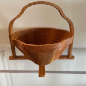 Puede incluir: Una cesta de madera hecha a mano con un asa curva y un diseño texturizado único. La cesta está hecha de madera de color marrón claro y presenta una forma distintiva, perfecta para uso decorativo o funcional.