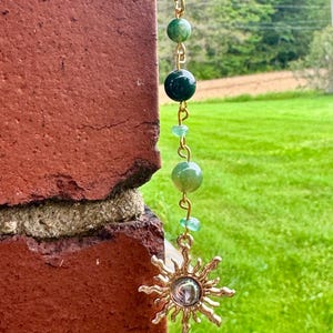 May include: A gold-toned ball chain necklace featuring a vertical arrangement of green beads in varying shades and sizes, connected by gold metal links, terminating in a gold-toned sun-shaped pendant with a central circular reflective element.