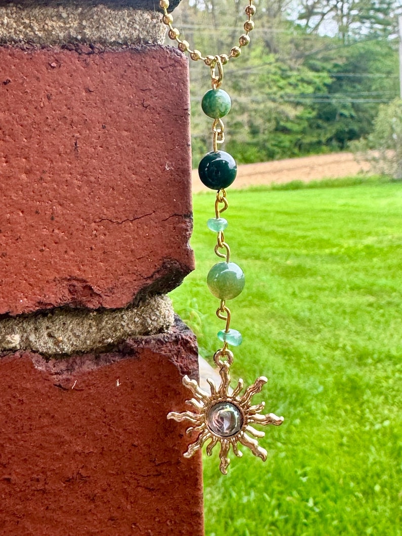 May include: A gold-toned ball chain necklace featuring a vertical arrangement of green beads in varying shades and sizes, connected by gold metal links, terminating in a gold-toned sun-shaped pendant with a central circular reflective element.