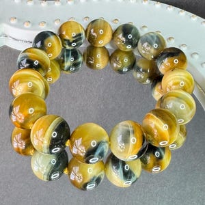 May include: A bracelet made of polished, round tiger eye beads. The beads are primarily golden-brown with dark brown and black stripes. The bracelet is strung on a gold-colored cord and is displayed on a white, decorative surface.