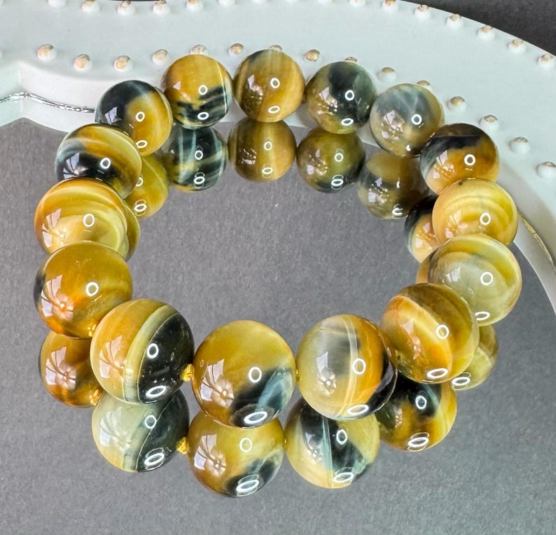 May include: A bracelet made of polished, round tiger eye beads. The beads are primarily golden-brown with dark brown and black stripes. The bracelet is strung on a gold-colored cord and is displayed on a white, decorative surface.