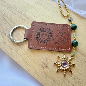May include: A brown rectangular leather keychain featuring a black engraved sun face and crystal design. It includes a silver metal ring and a gold-toned chain with green beads and a sun-shaped charm containing a central iridescent stone.