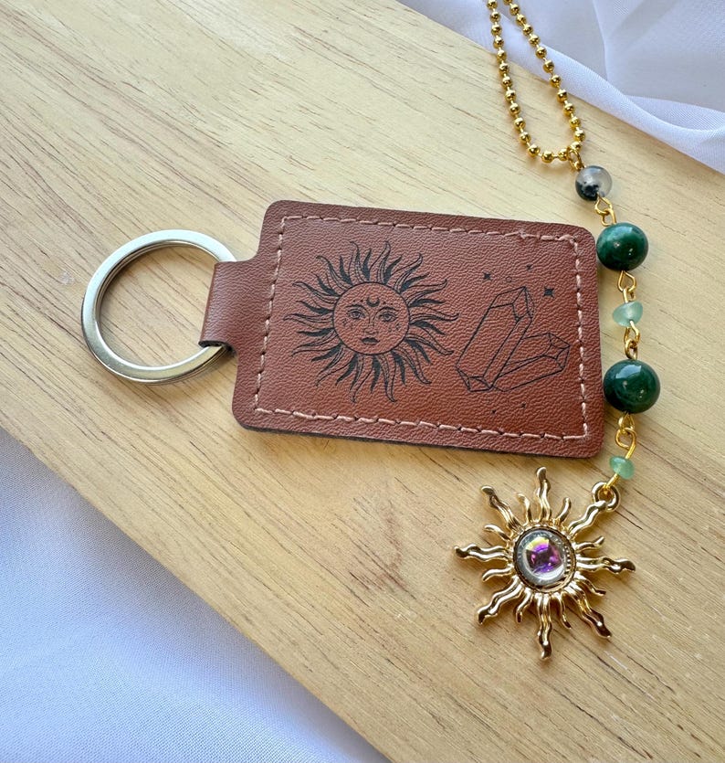 May include: A brown rectangular leather keychain featuring a black engraved sun face and crystal design. It includes a silver metal ring and a gold-toned chain with green beads and a sun-shaped charm containing a central iridescent stone.