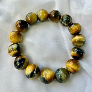 May include: A bracelet made of polished tiger's eye beads. The beads are a mix of golden yellow, brown, and dark blue, with a glossy finish. The bracelet is displayed on a white, textured surface.