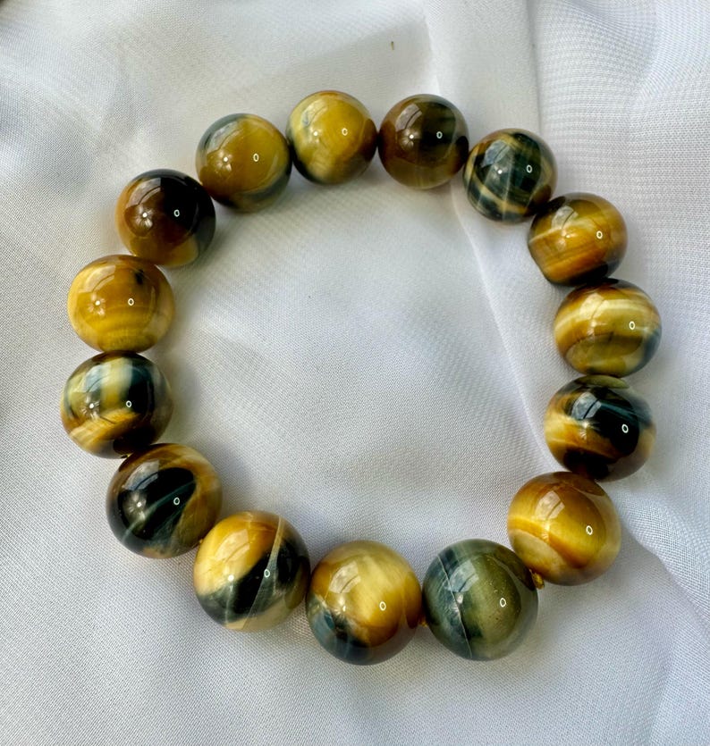 May include: A bracelet made of polished tiger's eye beads. The beads are a mix of golden yellow, brown, and dark blue, with a glossy finish. The bracelet is displayed on a white, textured surface.