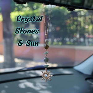 May include: A car hanging ornament featuring a gold-toned chain with alternating round and flat green and brown stones, ending in a gold-toned sun charm with a central clear crystal. The text Crystal Stones & Sun is visible on the left side.