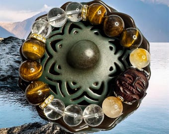 May include: A beaded bracelet featuring clear quartz, tiger's eye, and yellow gemstone beads. The bracelet is accented with gold-colored spacers and a carved wooden face. The bracelet is displayed on a decorative green ceramic plate.