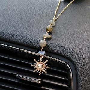 May include: A gold-toned chain car air freshener or decorative hanging ornament featuring a series of five smooth, multi-colored natural stones in shades of gray, green, and blue, ending in a gold-toned sun charm with a central white bead.