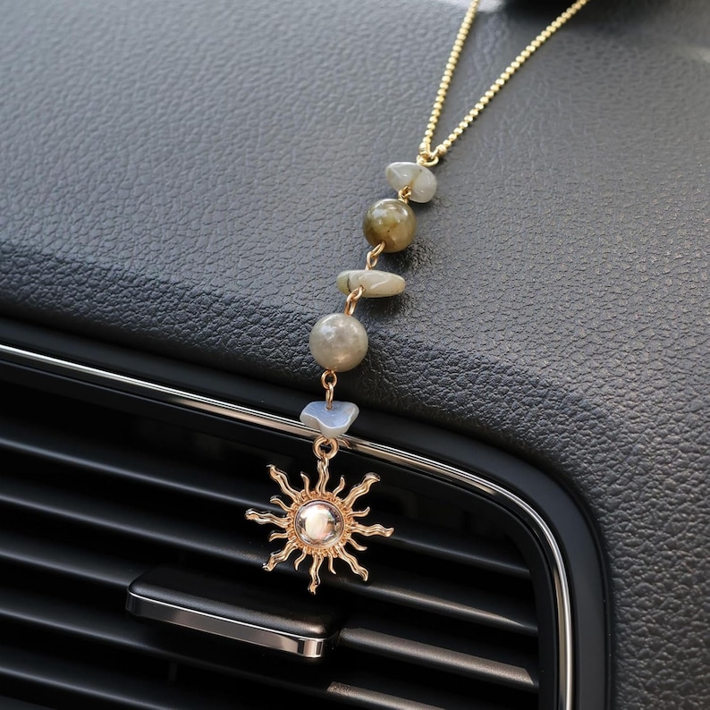 May include: A gold-toned chain car air freshener or decorative hanging ornament featuring a series of five smooth, multi-colored natural stones in shades of gray, green, and blue, ending in a gold-toned sun charm with a central white bead.