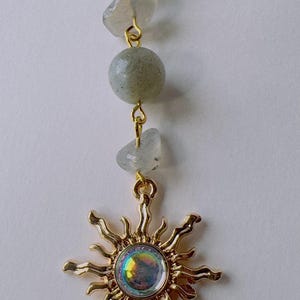 May include: A gold-toned sun pendant featuring a central iridescent circular stone with wavy rays, suspended from a vertical chain of alternating smooth round and irregular-shaped pale grey-green beads connected by gold-colored metal jump rings.