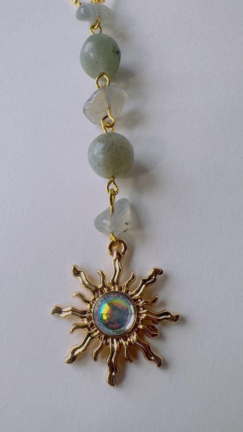 May include: A gold-toned sun pendant featuring a central iridescent circular stone with wavy rays, suspended from a vertical chain of alternating smooth round and irregular-shaped pale grey-green beads connected by gold-colored metal jump rings.