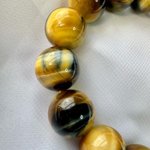 May include: Close-up of a bracelet featuring large, round beads. The beads have a polished, glossy finish with a mix of golden yellow, brown, and black colors. The bracelet is set against a white, textured background.