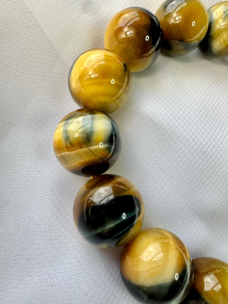 May include: Close-up of a bracelet featuring large, round beads. The beads have a polished, glossy finish with a mix of golden yellow, brown, and black colors. The bracelet is set against a white, textured background.