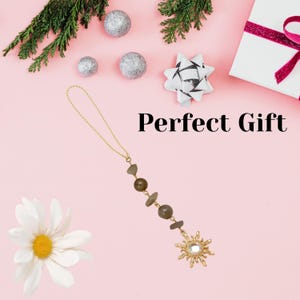 May include: A hanging ornament featuring a gold-toned chain, dark olive-green beads, and a sun-shaped charm with a central clear crystal. The text "Perfect Gift" is printed in black. The design includes a delicate gold loop for hanging purposes.