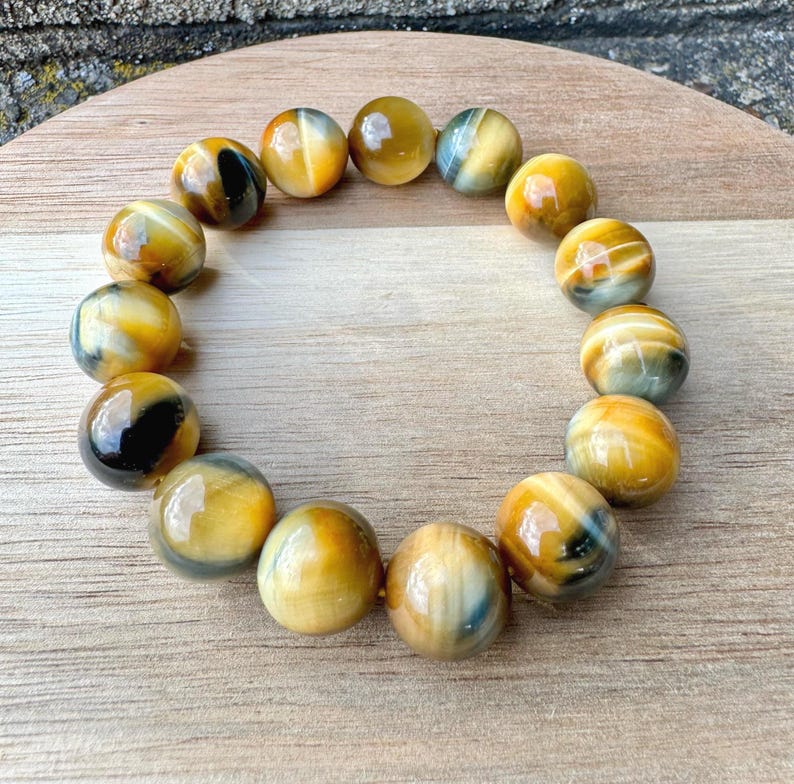May include: A bracelet made of polished, round tiger's eye beads. The beads are primarily golden yellow with streaks of black and blue. The bracelet is displayed on a wooden surface, showcasing the natural variations in the stone.