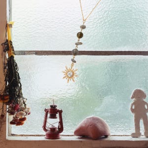 May include: A gold-toned necklace featuring a delicate chain with a vertical arrangement of three small grey stones and one larger olive-green bead, terminating in a sun-shaped pendant with a central clear stone set against a window.