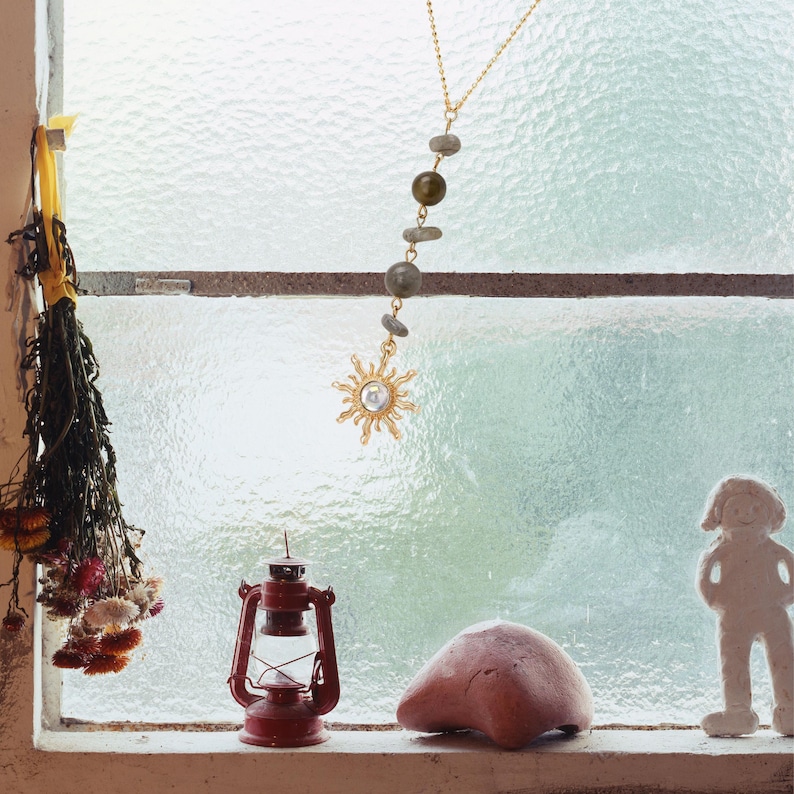 May include: A gold-toned necklace featuring a delicate chain with a vertical arrangement of three small grey stones and one larger olive-green bead, terminating in a sun-shaped pendant with a central clear stone set against a window.