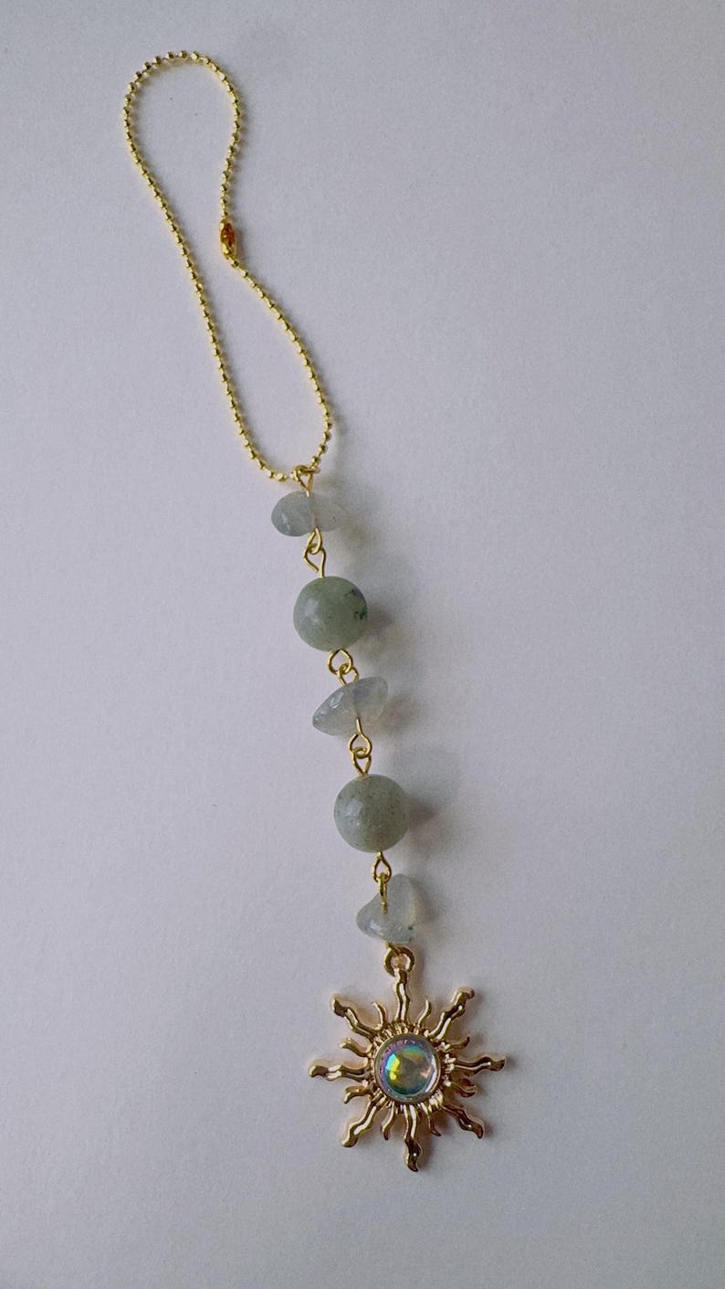 May include: A gold-toned ball chain ornament featuring a vertical arrangement of three pale blue-grey irregular stone chips and two smooth round sage green beads, terminating in a gold-toned sun charm with a central iridescent cabochon.