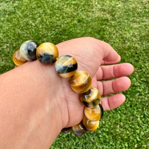 May include: A beaded bracelet featuring large, round tiger's eye stones. The stones display a mix of golden yellow, brown, and black hues. The bracelet is worn on a wrist against a backdrop of green grass.