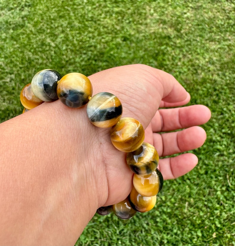 May include: A beaded bracelet featuring large, round tiger's eye stones. The stones display a mix of golden yellow, brown, and black hues. The bracelet is worn on a wrist against a backdrop of green grass.