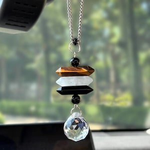 May include: A hanging car ornament featuring a silver-toned chain with three horizontal, double-pointed crystal beads in brown, white, and black, accented by two small black spherical beads and a clear, multifaceted spherical glass prism at the base.