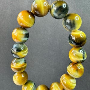 May include: A bracelet made of polished tiger's eye gemstone beads. The beads are a mix of golden yellow, brown, and blue-grey, strung together on a clear elastic cord. The spherical beads have a glossy finish.