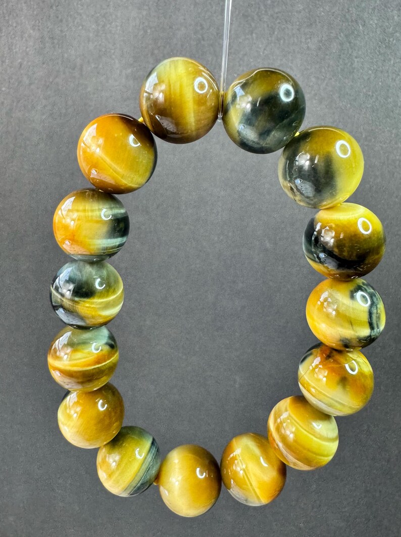 May include: A bracelet made of polished tiger's eye gemstone beads. The beads are a mix of golden yellow, brown, and blue-grey, strung together on a clear elastic cord. The spherical beads have a glossy finish.