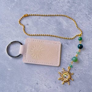 May include: A light pink rectangular keychain with an embossed sun and crystal design, attached to a gold-toned ball chain. The chain features green beads and a gold-toned sun charm with a central iridescent stone, finished with a metal ring.