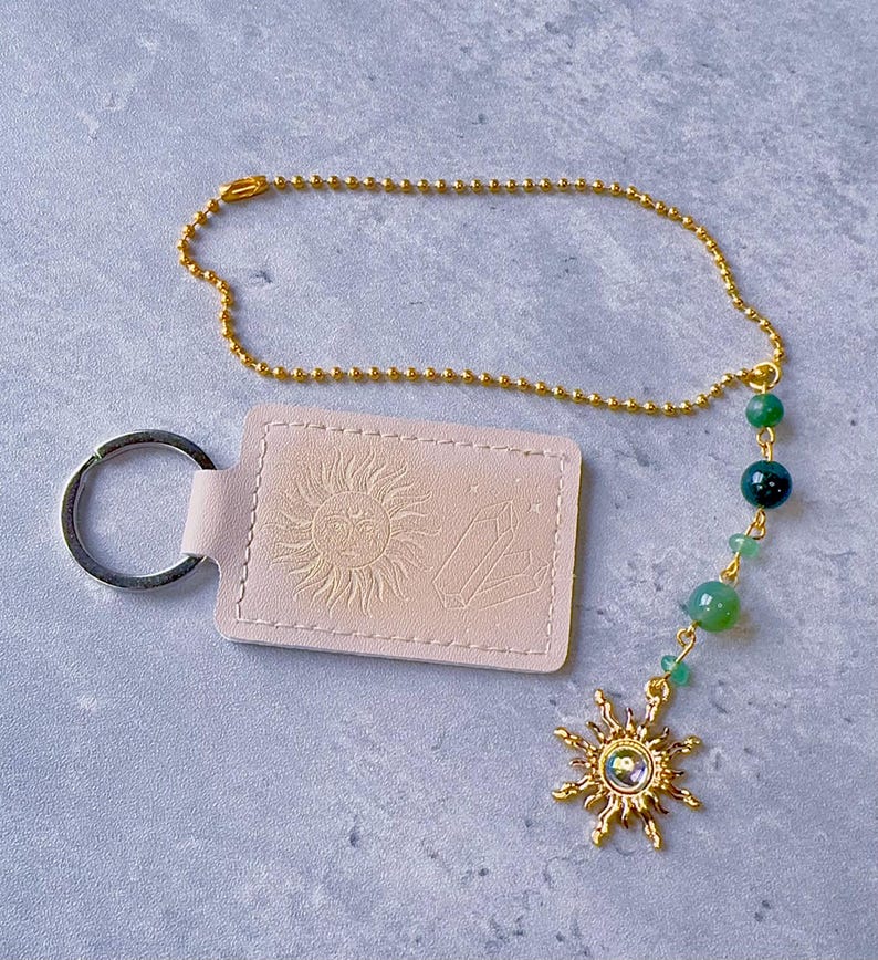 May include: A light pink rectangular keychain with an embossed sun and crystal design, attached to a gold-toned ball chain. The chain features green beads and a gold-toned sun charm with a central iridescent stone, finished with a metal ring.