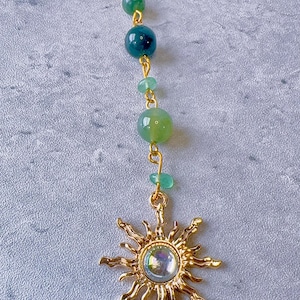 May include: A gold-toned ball chain necklace featuring a vertical arrangement of green beads in varying shades and sizes, terminating in a gold-toned sun-shaped pendant with a central iridescent cabochon stone set in the middle of the sun rays.