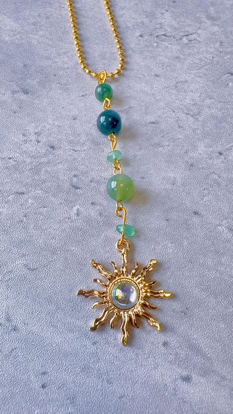May include: A gold-toned ball chain necklace featuring a vertical arrangement of green beads in varying shades and sizes, terminating in a gold-toned sun-shaped pendant with a central iridescent cabochon stone set in the middle of the sun rays.