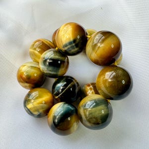 May include: A bracelet featuring large, polished tiger's eye beads. The beads display a mix of golden brown and deep blue-green hues, strung together on a yellow cord. The spherical beads have a glossy finish.